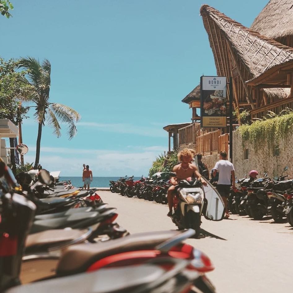 a man driving his scooter past a row of scooters and shops near Berawa Beach